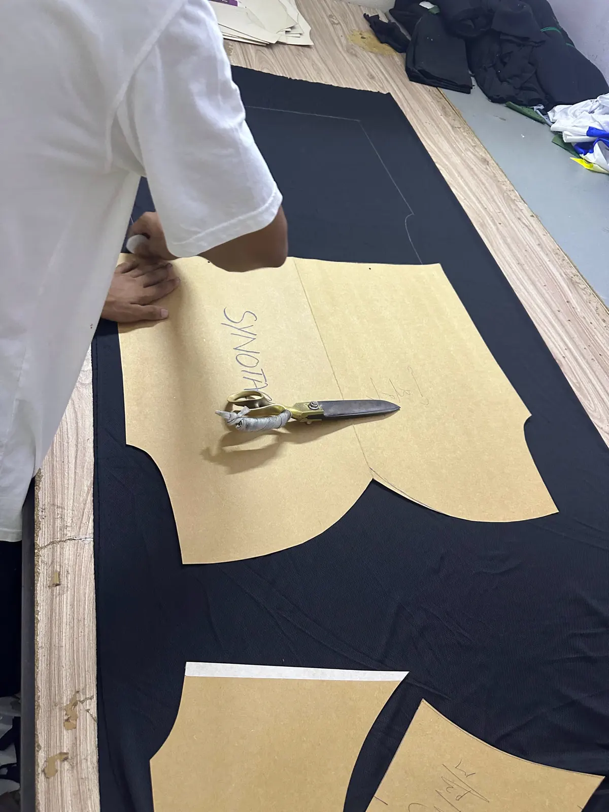 Brown paper pattern pieces, one marked Synotacy, laid over black fabric on a cutting table, with tailor's shears resting on top and a hand smoothing the pattern.