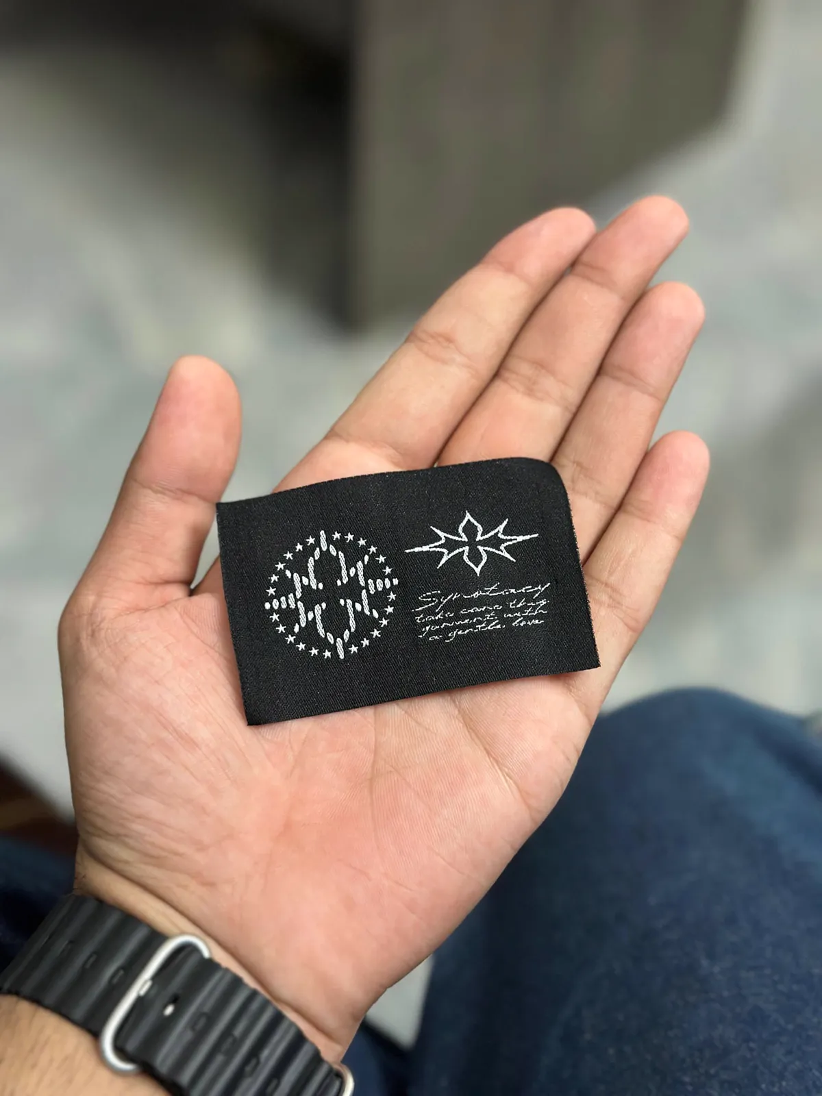 An open palm holding a black woven Synotacy label with a starred monogram roundel, the star mark and a handwritten care line.