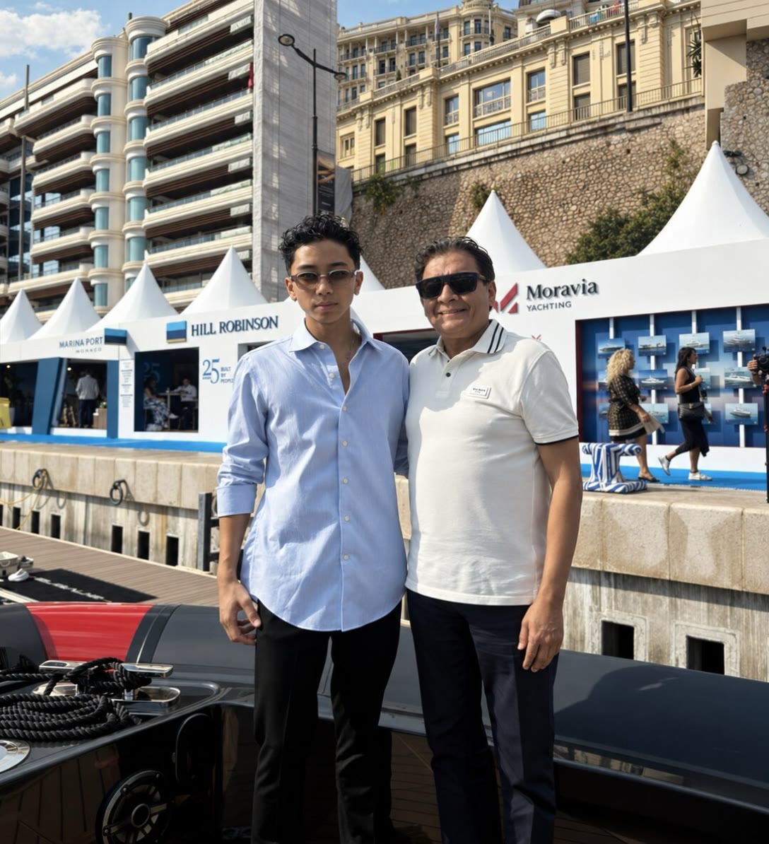 Ryan Capistrano and Sean Rashid stand together on a boat deck at the Monaco Yacht Show, exhibitor tents and harbour buildings behind them.
