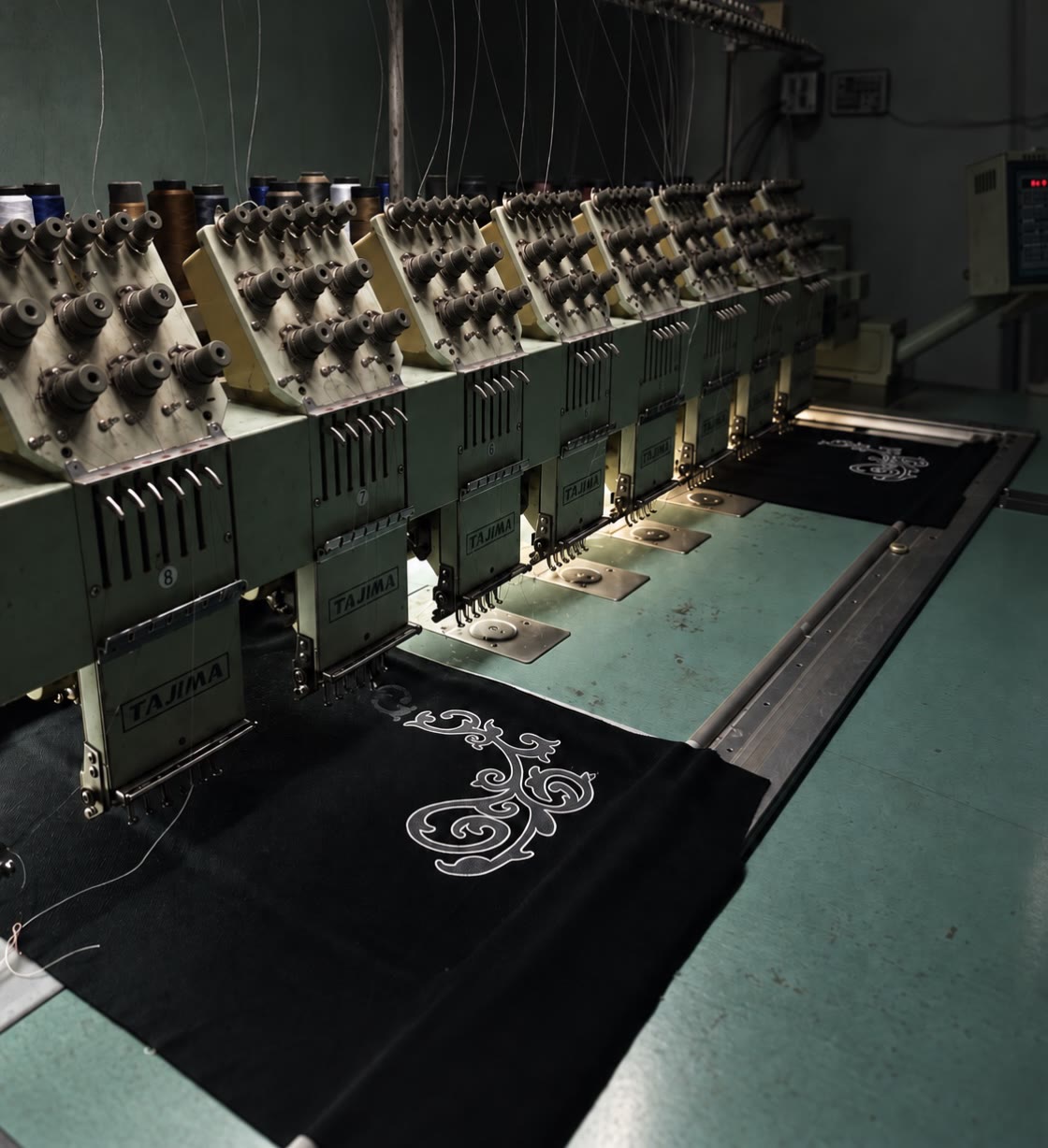 A row of industrial multi-head embroidery machines stitching a white ornament onto black Synotacy panels.