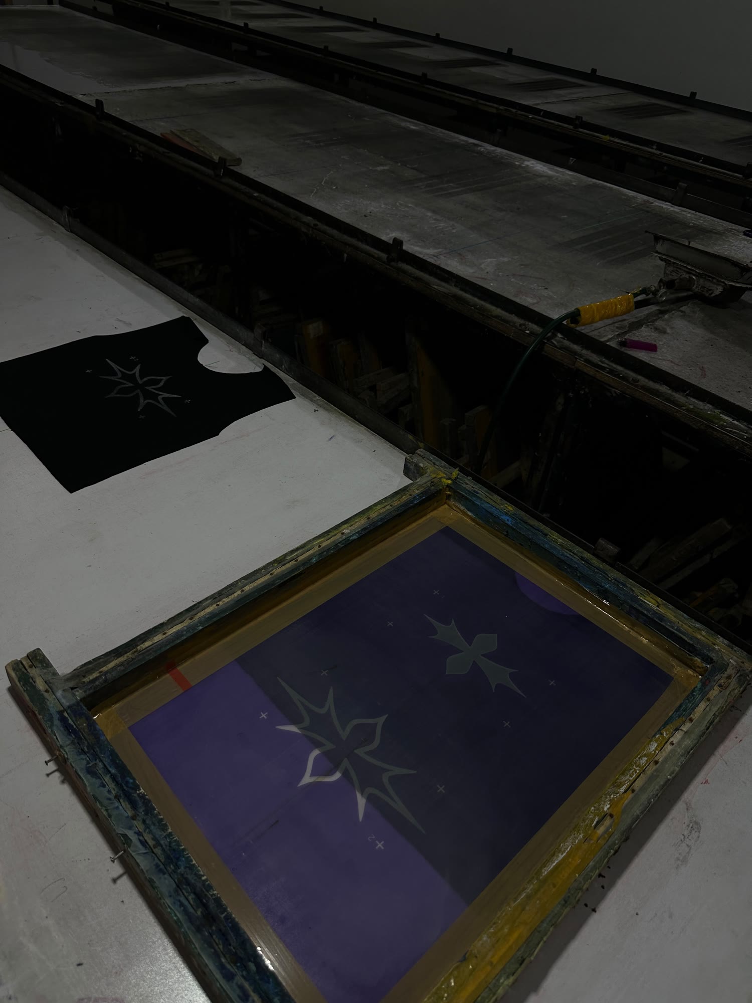 A wooden screen-printing frame on a long print table, the Synotacy star mark burned into the violet mesh, a printed black panel behind it.