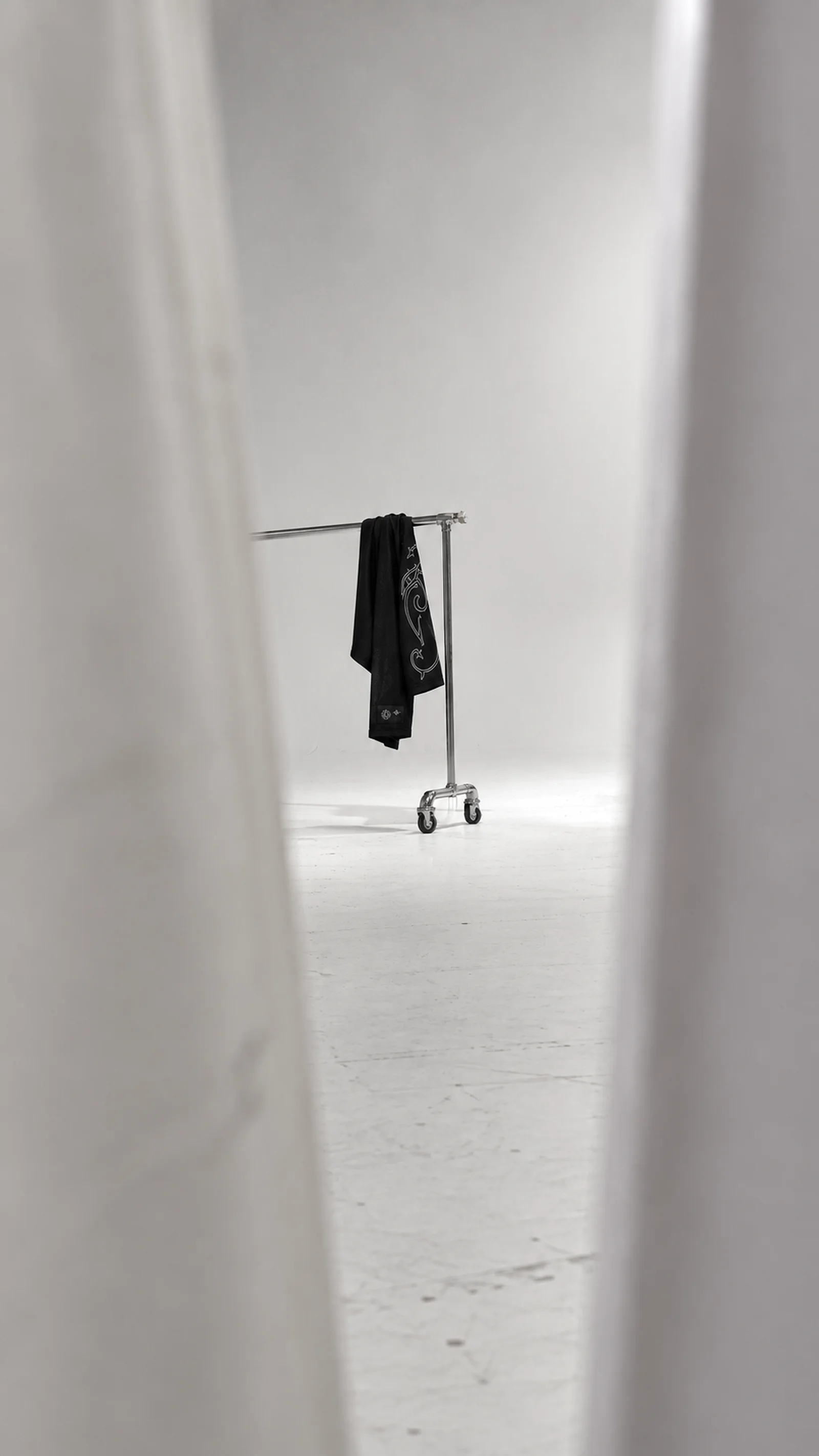 One black garment with a pale embroidered ornament hangs on a chrome rolling rack in a white studio, framed by the gap between two white curtains.